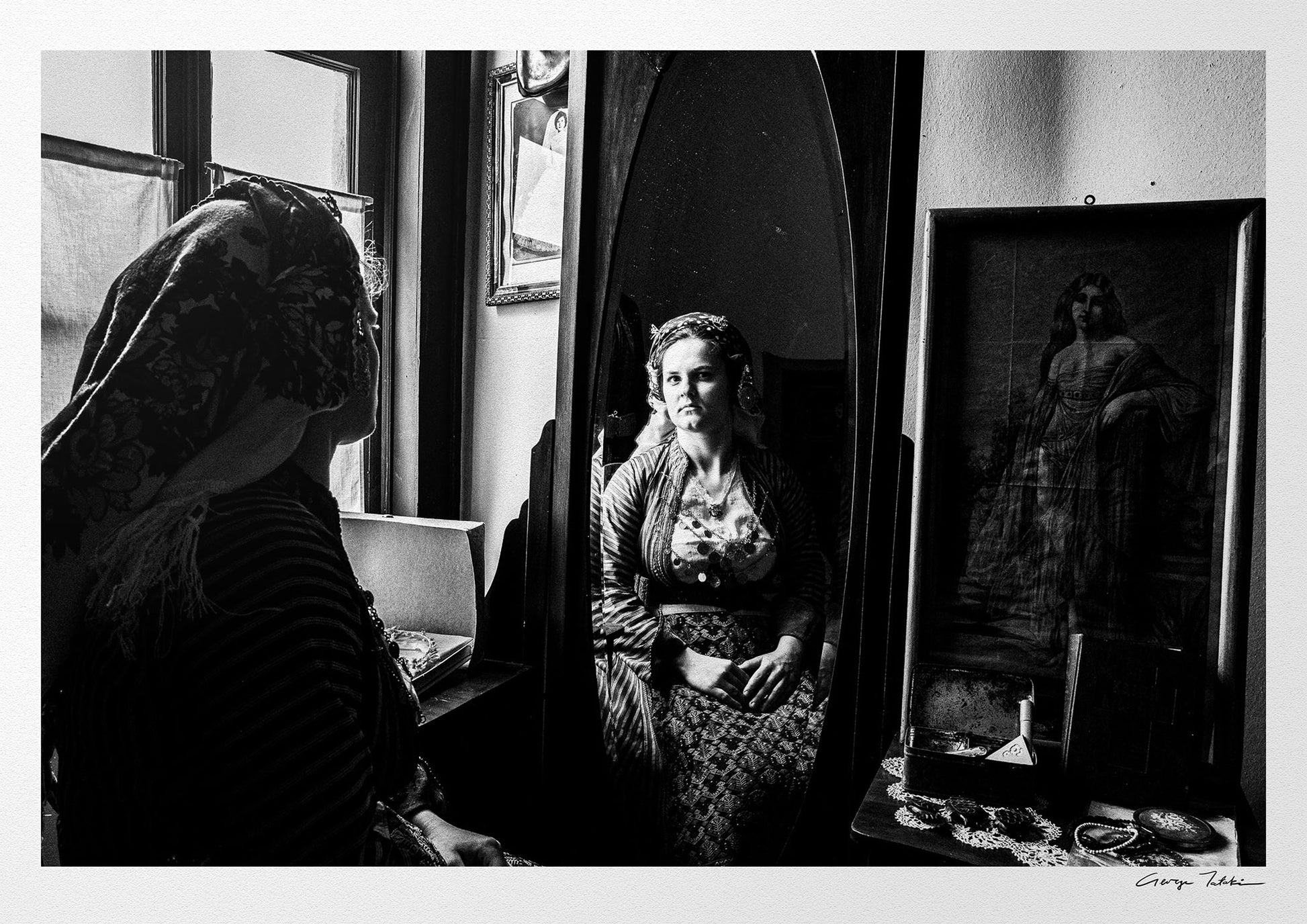 Woman looking at herself in a mirror in Soufli, Thrace, Greece, with a vintage photograph on the wall. Black and white photography by George Tatakis