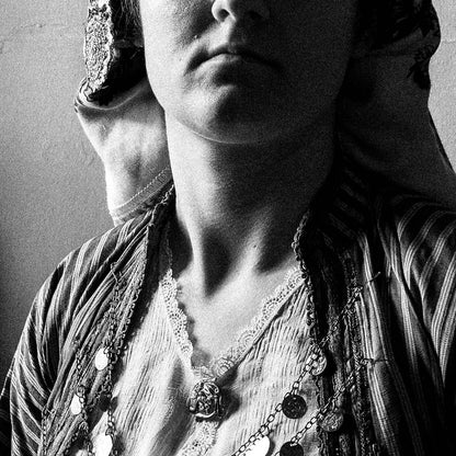 Black and white portrait of a woman in a room with framed pictures on the wall wearing the bridal dress of Soufli, Thrace, Greece. Photography close-up detail by George Tatakis