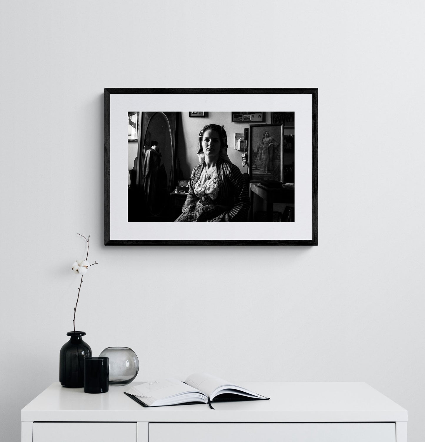 Black and white portrait of a woman in a room with framed pictures on the wall wearing the bridal dress of Soufli, Thrace, Greece. Photography framed on a wall by George Tatakis