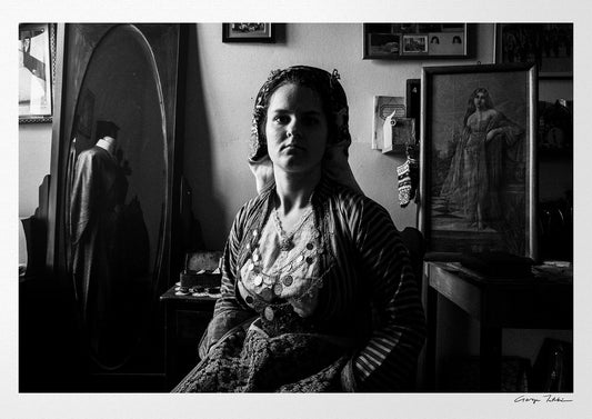 Black and white portrait of a woman in a room with framed pictures on the wall wearing the bridal dress of Soufli, Thrace, Greece. Photography by George Tatakis