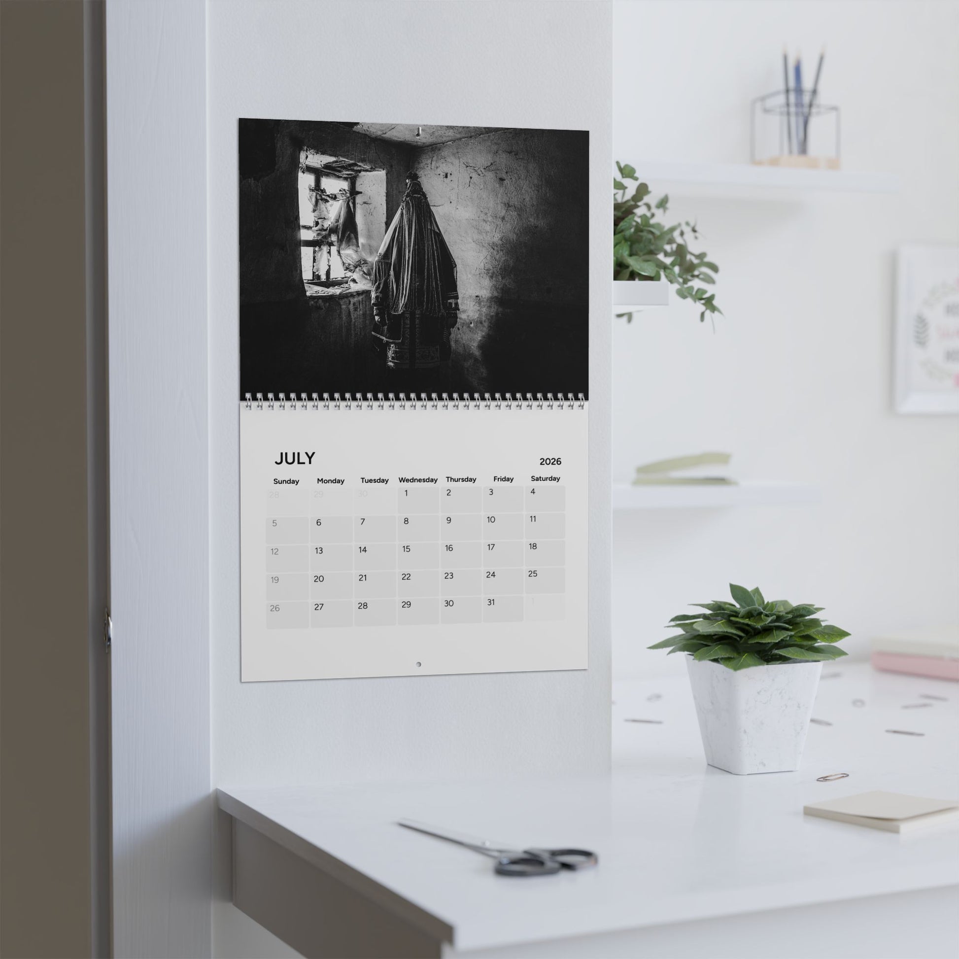 Calendar on a wall with a black and white photo of a person hanging clothes, in a room with a desk and plant.