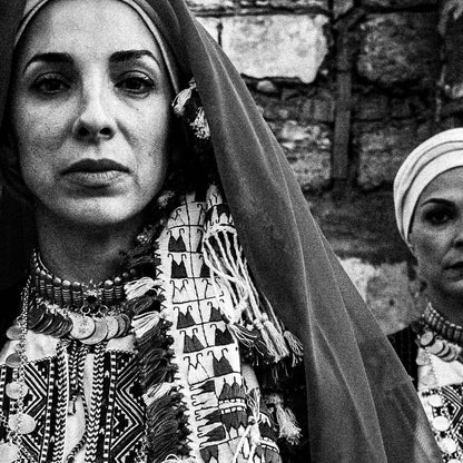 Unveiled bride in traditional attire standing in front of a stone wall. Kyparissia, Peloponnese, Greece. Black and white photography close-up detail by George Tatakis.