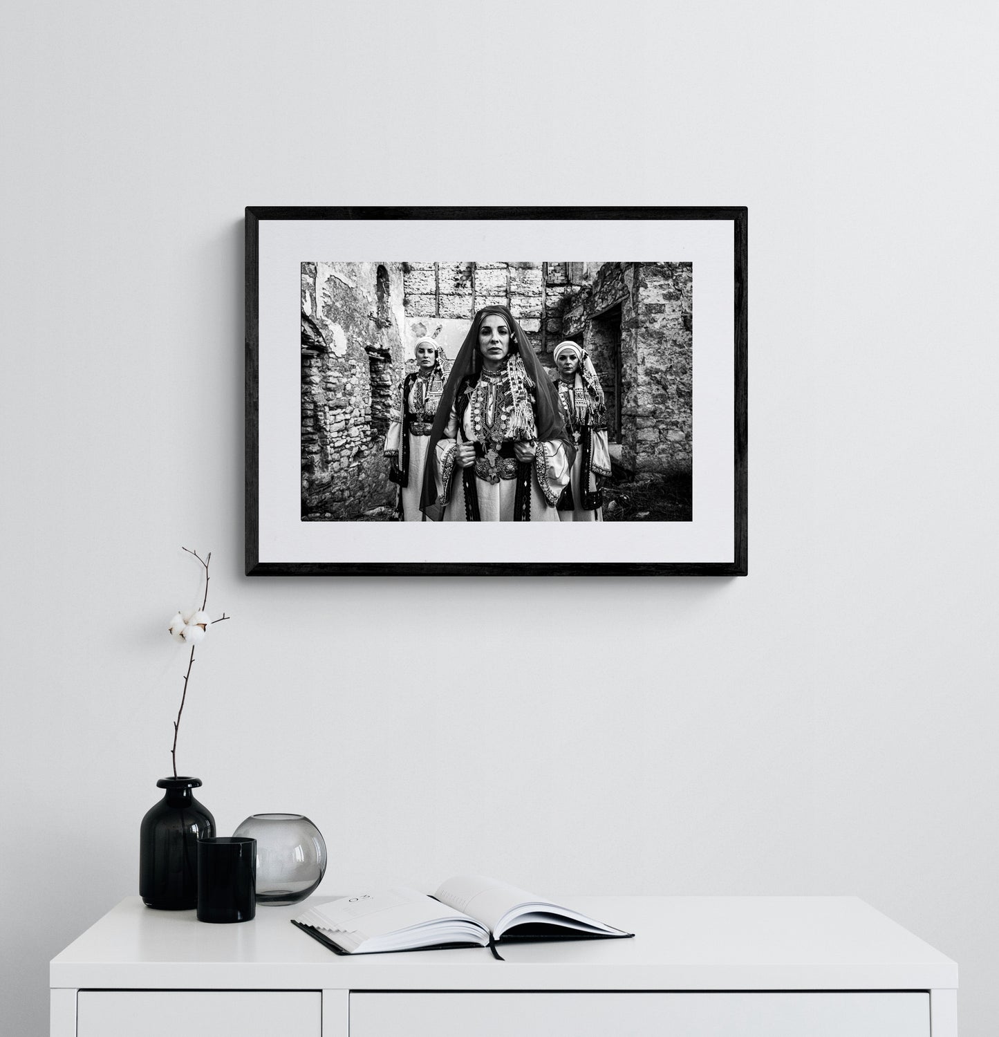 Unveiled bride in traditional attire standing in front of a stone wall. Kyparissia, Peloponnese, Greece. Black and white photography framed on a wall by George Tatakis.