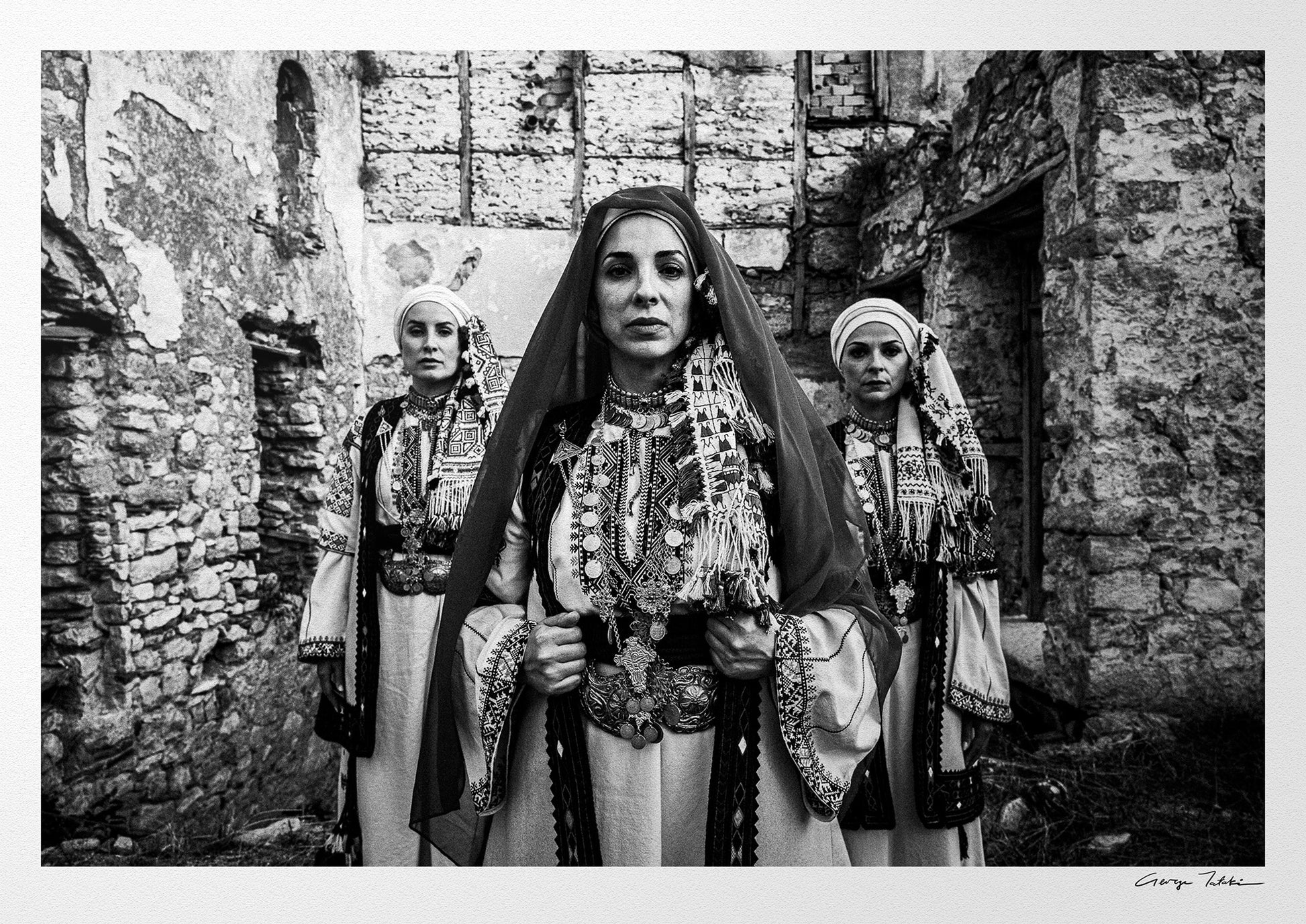 Unveiled bride in traditional attire standing in front of a stone wall. Kyparissia, Peloponnese, Greece. Black and white photography by George Tatakis.