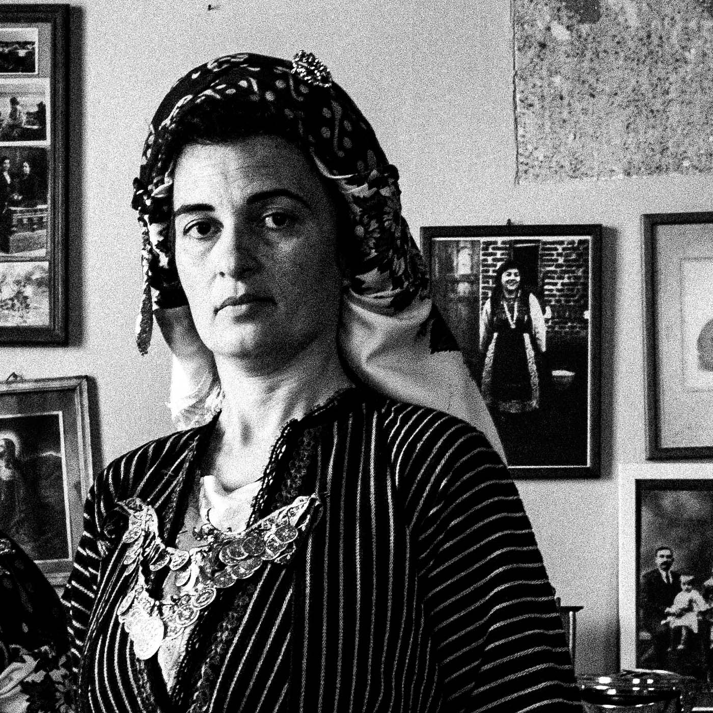 Two women in a room with framed pictures on the wall. Local dresses of Soufli, Thrace, Greece. Black and white photography close-up detail by George Tatakis.