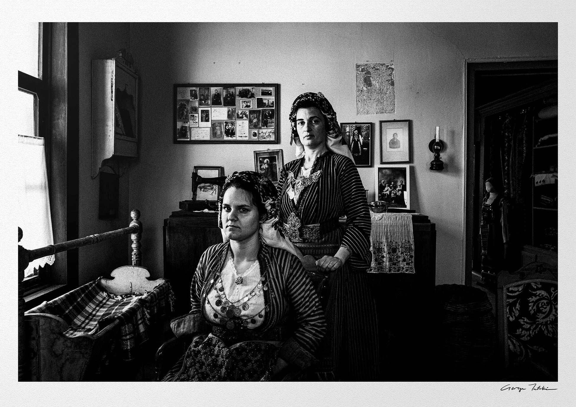 Two women in a room with framed pictures on the wall. Local dresses of Soufli, Thrace, Greece. Black and white photography by George Tatakis.