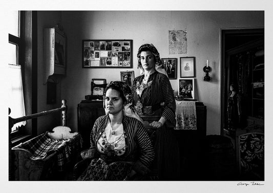Two women in a room with framed pictures on the wall. Local dresses of Soufli, Thrace, Greece. Black and white photography by George Tatakis.